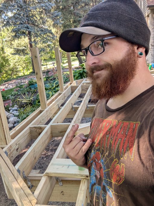 A bearded man wearing glasses, a cap, and a Marvel 'Daredevil' t-shirt pointing toward a wooden raised garden bed frame in his backyard.