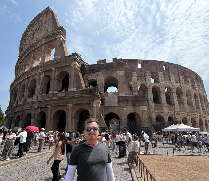 A man poses in front of the iconic Colosseum in Rome on a bright, sunny day surrounded by tourists. The caption "bullish" playfully nods to the Colosseum's history of gladiatorial combat, where bulls and fighters battled—evoking strength, power, and a fighting spirit.