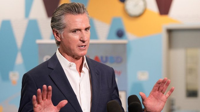 A man in a dark blazer speaks with raised hands in front of microphones, with partial "UB" text visible in the background.

The image appears to show California Governor Gavin Newsom, who is referenced in the post as having signed AB 2409 into law, banning public officials in California from issuing memecoins.