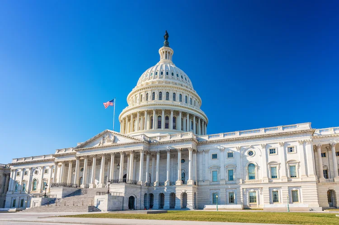 A clear daytime photograph of the United States Capitol building in Washington D.C., featuring its iconic white dome and an American flag flying against a blue sky.