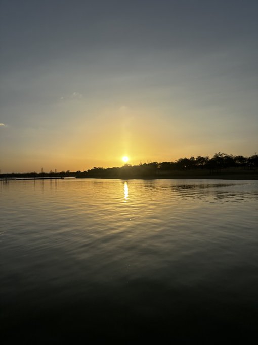 A scenic photograph of a sunset over water, with the sun reflecting on the calm surface and a silhouette of trees and land on the horizon.