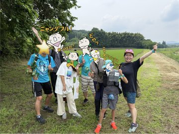 The image features a group of people posing for a photo, with some holding up their heads as if they are cartoon characters or superheroes. The group consists of at least seven individuals standing together in front of trees, giving off an impression that they might be part of a comic book or superhero team. They appear to be enjoying themselves and having fun during the photo session.