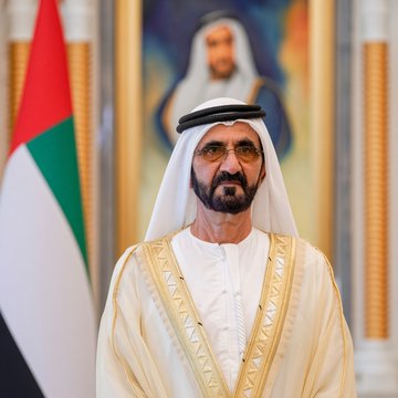 A man in traditional white Gulf Arab attire stands before a UAE flag, wearing glasses and a beard.