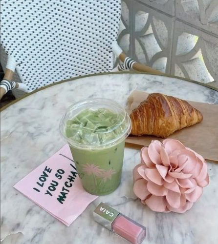 A pink matcha latte, a croissant, a Chanel lipstick, a pink flower, and a napkin reading "I LOVE YOU SO MUCH" sit on a marble table.