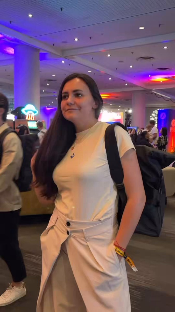 The image features a woman standing in an airport terminal, wearing a white shirt and beige pants with her hair pulled back into a ponytail. She has a backpack on her shoulders and appears to be posing for a photo. The background of the image shows other people walking around the terminal, indicating that it is a busy location. There are also signs visible in the scene, which might provide information about the airport or its services.