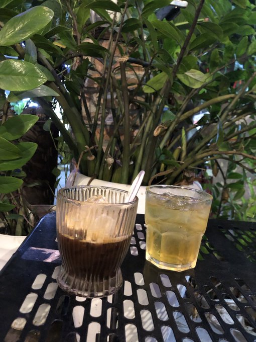 A casual photo of a coffee in a ribbed glass and a glass of iced tea with a spoon, sitting on a black perforated table with lush green plants in the background.