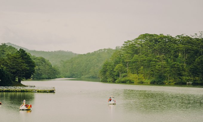 A serene lake scene with people on swan-shaped paddle boats, surrounded by lush green forested hills under a hazy sky.
