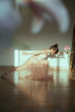 An artistic photograph of a ballerina in a pink tutu stretching on a wooden floor, with blurry flower petals floating in the foreground, bathed in soft light.