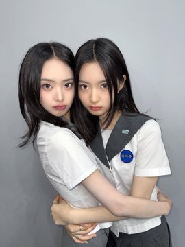 Two young women with long dark hair embrace each other in matching white shirts, one wearing a NASA badge, posing closely together against a plain gray background.