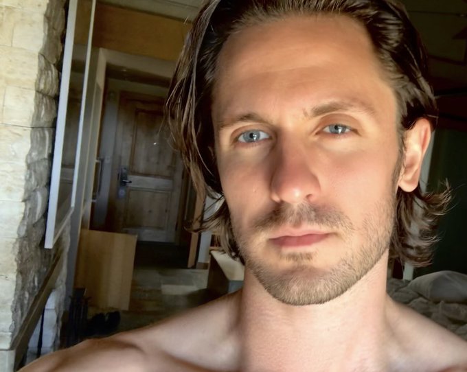 A shirtless man with long brown hair and blue eyes takes a close-up selfie, appearing relaxed as he reflects humorously on missing an early investment opportunity. The casual setting and his somewhat bemused expression mirror the "facepalm" tone of lamenting a missed $BAYLA investment in the Jungle BayAC NFT project.