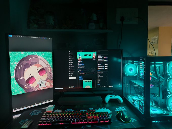 A dimly lit gaming setup features dual monitors displaying the name "IRYS" on a chat interface, alongside an anime character wallpaper and a RGB-lit PC with an Xbox controller, creating a cozy setup that matches the "Irys is love" appreciation shown in the post.