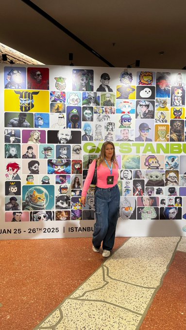 A woman in a pink top and denim overalls poses in front of a colorful wall covered with profile avatars at a gaming convention, with partial text reading 'GAM... STANBUL' and 'JAN 25 - 26TH 2025 | ISTAN...'.