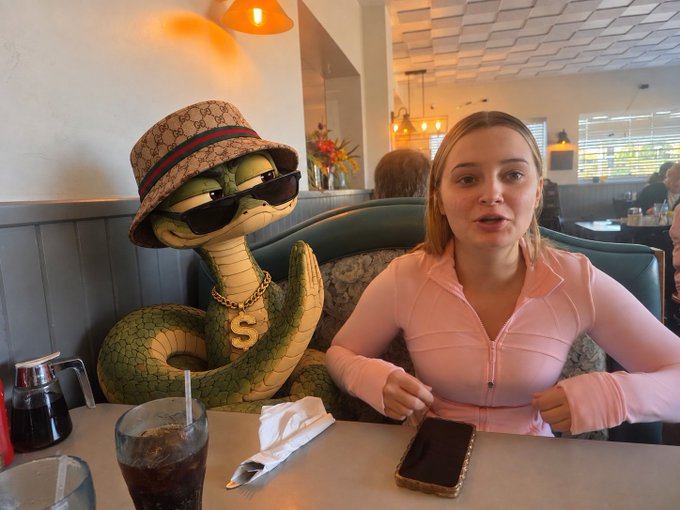 A young woman in a pink zip-up top sits at a restaurant table next to a cartoonish statue of a snake wearing sunglasses, a Gucci bucket hat, and a gold chain with a dollar sign pendant.