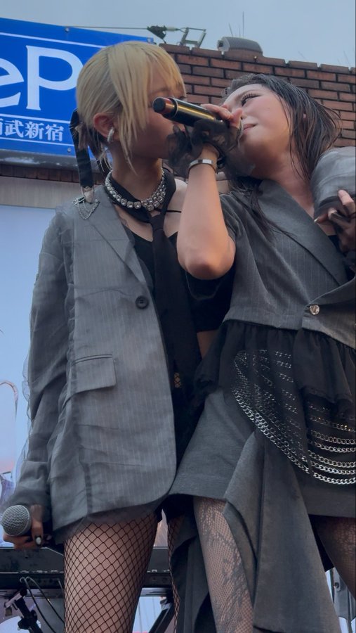Two performers on an outdoor stage, one with blonde hair in a grey blazer and the other in a black outfit with chain details, share a microphone.