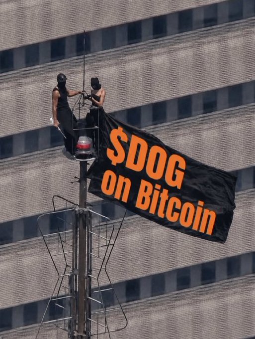 Two people in black tank tops and caps stand on a high tower structure, holding a black flag with orange text reading '$DOG on Bitcoin', referencing the Dogecoin cryptocurrency.