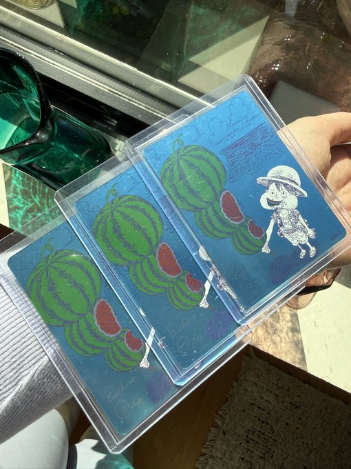A hand holds three acrylic charms featuring watermelon illustrations in progress, including a character wearing a hat, labeled 'Enchanto', with sunglasses resting nearby.