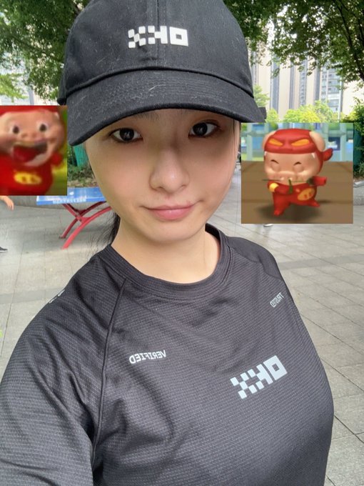 A selfie of a young woman wearing a black 'OKX' branded cap and matching shirt, with two small cartoon pig/monkey character stickers overlaid on the sides.