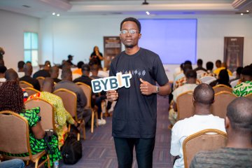 The image shows a man standing in front of an audience at a conference or event. He is holding up a sign that says "BYBT" while giving a thumbs-up gesture, indicating his enthusiasm for the topic being discussed. The room contains several chairs arranged around the space and people sitting on them, suggesting it's a gathering place for attendees to listen and engage in conversation.