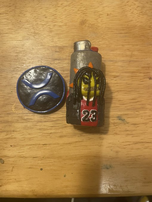 A lighter featuring a small figure wearing a red '23' jersey sits on a wooden table next to a round metal object with a blue swirl design.