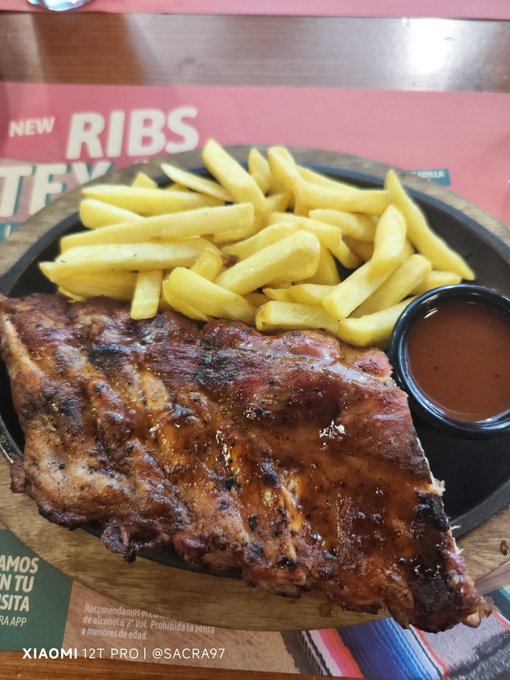 A plate of BBQ ribs with golden french fries and a side of barbecue sauce, sitting on a restaurant table with a menu promoting 'NEW RIBS TEX', captured by Xiaomi 12T Pro.