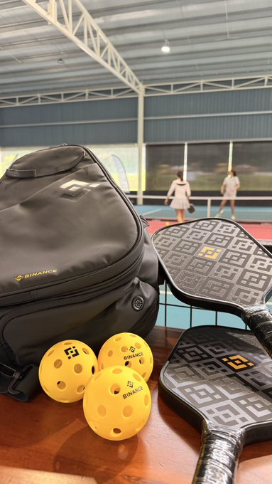 A sports bag and pickleball paddles branded with the "BINANCE" logo sit on a wooden table with yellow balls, overlooking an indoor sports court.