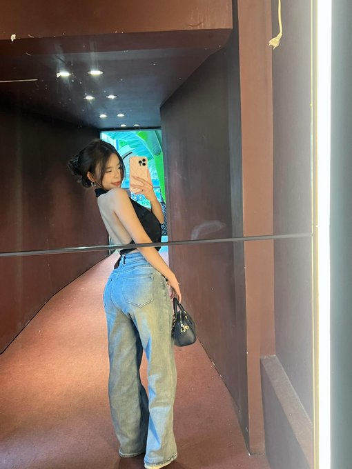 A woman poses for a mirror selfie in a hallway, wearing a black halter top, blue jeans, and holding a small black handbag, with the reflection captured in what appears to be a glass surface.
