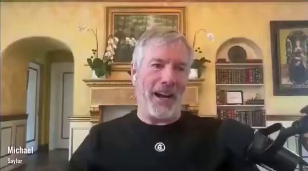 A man sitting behind a microphone with his hand over his mouth saying Michael said they plan to buy $70,000,000,000 worth of Bitcoin driving the price to $10 million.