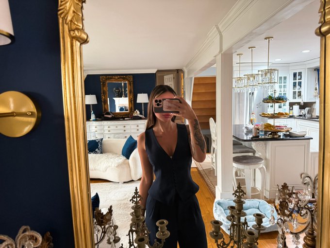 A woman in a navy blue peplum outfit takes a mirror selfie in an elegant home interior featuring a gold-framed mirror, ornate chandelier, and classic decor.