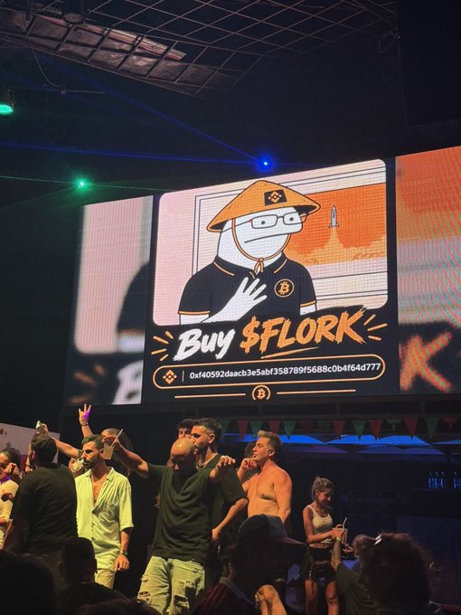 A crowd gathers at what appears to be a club or event in Thailand, where a large screen displays the "Buy $FLORK" meme featuring a cartoon character in an orange hat alongside a crypto wallet address (0xf40592daacb3e5abf358789f5688c0b4f64d777), directly supporting the post about spreading the $FLORK token in Thailand.