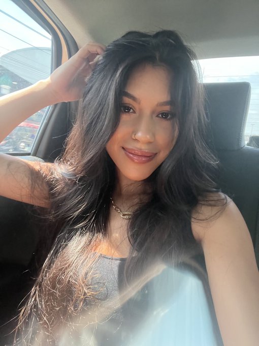 Selfie of a young woman with long dark hair sitting in a car, wearing a gray top and a gold necklace.