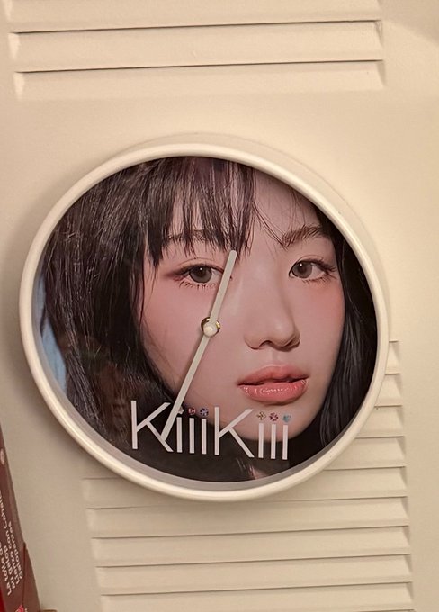 A circular clock or device featuring a portrait of a young woman with dark hair and bangs, with the text 'KiiiKiii' displayed across the bottom.