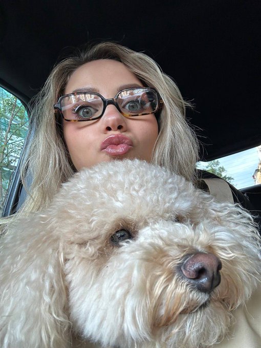 A selfie taken inside a car featuring a woman with glasses making a kissing face next to a fluffy, cream-colored dog.