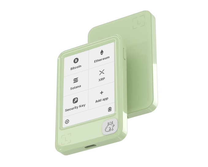 A pair of pale green hardware crypto wallets are displayed, with one showing a screen listing "Bitcoin," "Ethereum," "XRP," "Solana," "Security Key," and "Add app." The "green is all around you" caption reflects the device's fresh mint-green casing, tying the eco-friendly aesthetic to the crypto security device.