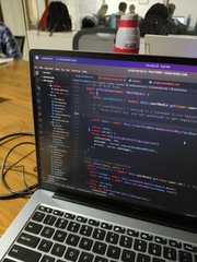 A photograph of a laptop displaying lines of code in a dark-themed editor, with a water bottle visible behind the screen in what appears to be an office or classroom setting.
