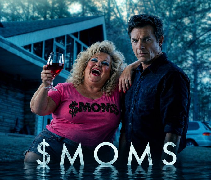 A movie or TV poster titled '$MOMS' features a laughing blonde woman in a pink '$MOMS' shirt holding a glass of wine, standing next to a serious-looking man in a dark shirt, with the title reflected in water below them.