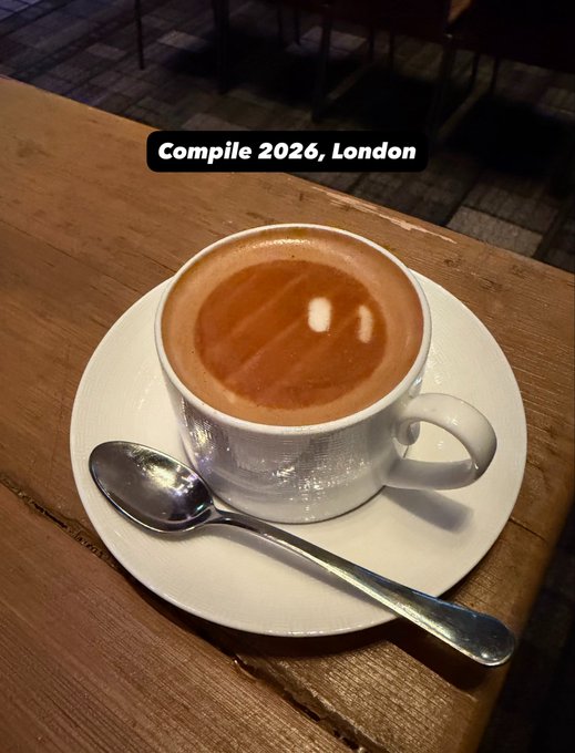 A steaming cup of coffee sits on a white saucer with a spoon, accompanied by the text "Compile 2026, London" — capturing the cozy, early-morning coffee moment shared with the "Good Morning from Compile London!" greeting.