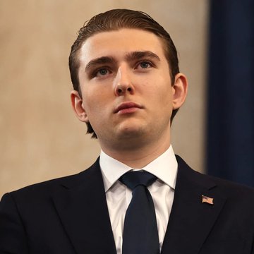 Barron Trump, dressed in a navy suit with an American flag lapel pin, looks off-camera in this close-up portrait. The image accompanies reports of his brief social media post endorsing Bitcoin, which he deleted within 20 seconds of posting.
