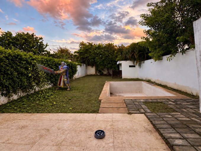 A character in armor holding a large sword stands in a grassy backyard at sunset, near a concrete pit and a white wall.