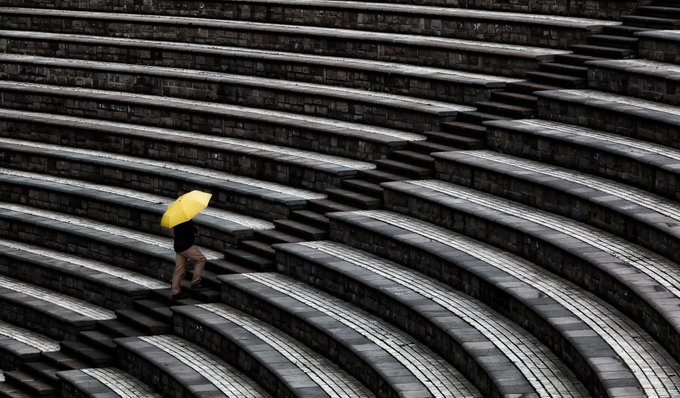 A person holding a yellow umbrella walks alone across the curved, empty stone steps of an amphitheater.