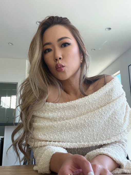 A woman with long wavy blonde-highlighted hair takes a selfie in a cream off-shoulder sweater, pursing her lips playfully while leaning forward with a sassy expression in a bright modern interior. The image appears unrelated to the post's critique of misaligned crypto and tech marketing budgets, serving instead as an attention-grabbing personal photo to draw engagement to the commentary about inflated marketing spending.