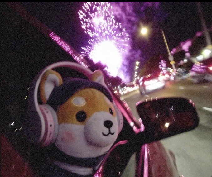 A Shiba Inu plush toy wearing pink headphones sits in a car, watching a fireworks display light up the night sky through the window, with the side mirror reflecting city lights.