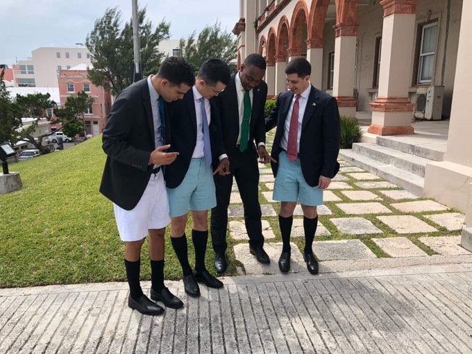 Four men in formal blazers, ties, and knee-high socks pose together while wearing light blue and white Bermuda shorts, illustrating a playful take on traditional Bermuda attire. The scene captures a lighthearted moment of "Bermuda Culture" with CZ's official Bermuda shorts front and center.