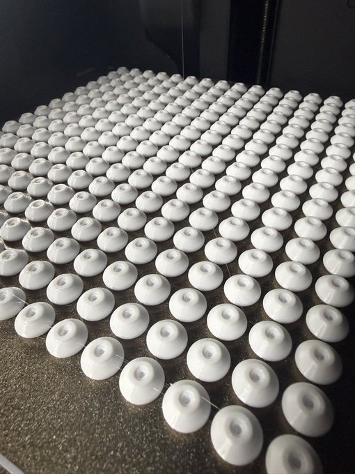 A large array of small white circular 3D-printed objects arranged in neat rows on a dark surface, each featuring a central hole and subtle markings.