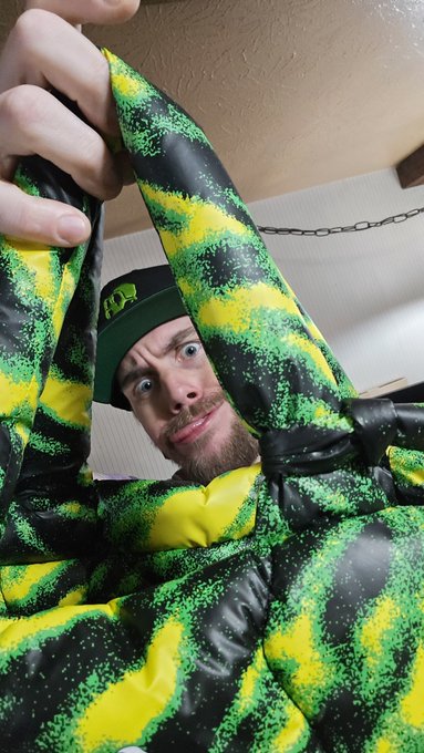 A selfie of a bearded man wearing a green cap, peering through the opening of a black and yellow patterned puffer jacket.
