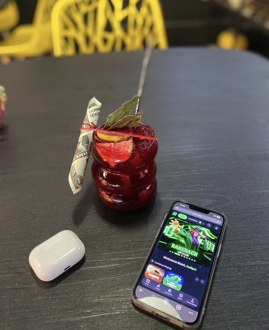 A dark table holds an AirPods case, a smartphone displaying a gaming app called 'RAKEBACK', and a red decorative item with a dollar bill attached.