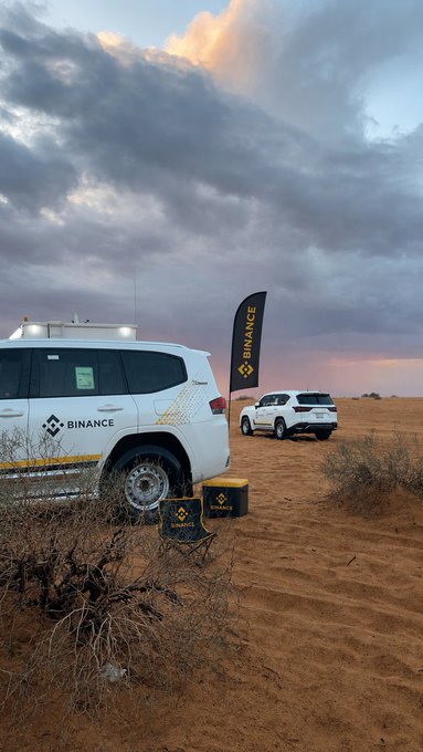 The image shows a white car parked next to two other cars in a desert setting. There are also two small boxes placed near the vehicles. The backdrop features a cloudy sky, which complements the overall scene. The text "From the city's streetlights to the dryness of the desert, always with Binance" is present, suggesting that the image might be related to Bitcoin or cryptocurrency in some way.