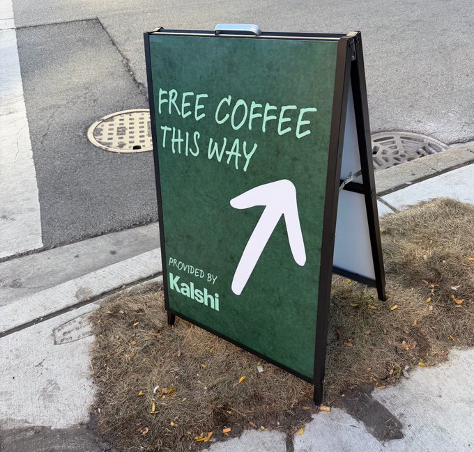 An A-frame street sign on a patch of grass and dirt that reads 'FREE COFFEE THIS WAY' with an arrow pointing diagonally, and 'PROVIDED BY Kalshi' in the corner.