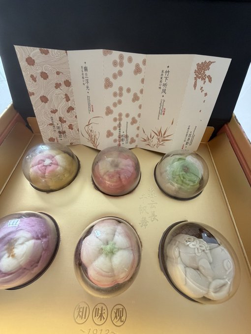 A gold gift box displays six intricately designed mooncakes shaped like peonies and lotuses in pastel pink, green, and white, accompanied by decorative cards reading "竹下听风" and "如玉生花" alongside the brand name "知味观 (1913)." The image supports the post's celebration of Mid-Autumn Festival by showcasing the elegant mooncake gift set being given away in the giveaway.