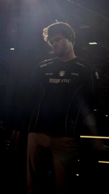 A man with curly hair stands in a dimly lit, atmospheric venue wearing a black racing-style jersey featuring 'imc' sponsor logos.