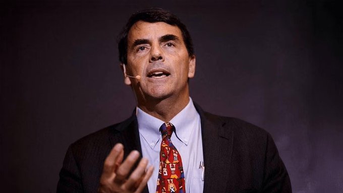 A man in a dark suit, light blue shirt, and colorful patterned tie speaks into a headset microphone while gesturing with his hand, captured mid-speech against a dark purple background. The image illustrates the accompanying post about Tim Draper's bold Bitcoin price prediction regarding its value against the US dollar.